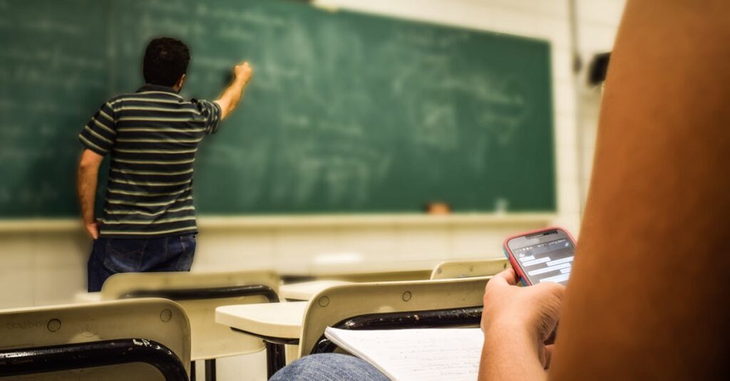 Student texting in a classroom while teacher is writing on the blackboard.